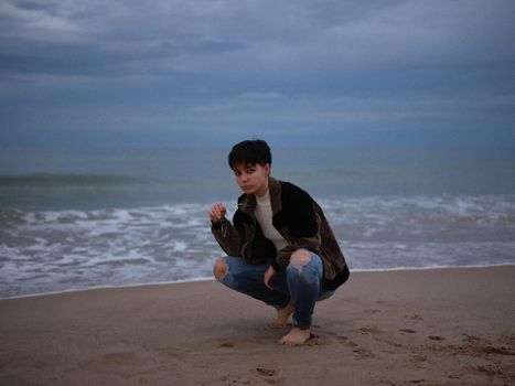 trans woman squatting with the beach behind her, posing in warm clothes, cloudy day