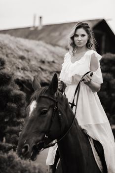 A woman in a white sundress riding a horse near a farm. black and white photo.