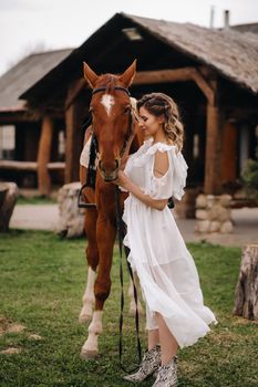 Beautiful girl in a white sundress next to a horse on an old ranch.