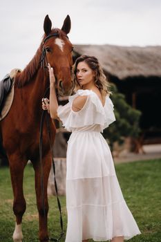 Beautiful girl in a white sundress next to a horse on an old ranch.