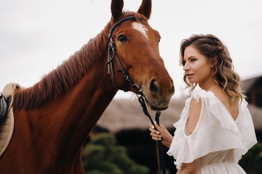 Beautiful girl in a white sundress next to a horse on an old ranch.