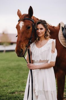 Beautiful girl in a white sundress next to a horse on an old ranch.