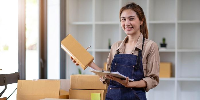 Young attractive asian woman owner startup business look at camera work happy with box at home prepare parcel delivery in sme supply chain..