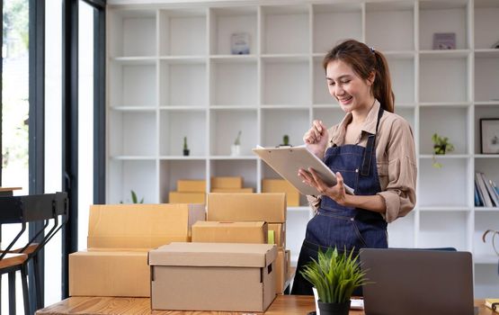 Beautiful smart Asian young entrepreneur business woman owner of SME checking product on stock and write on clipboard working at home. Small business owner at home office concept..