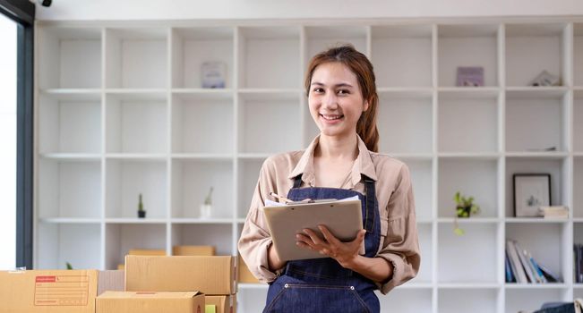 Beautiful smart Asian young entrepreneur business woman owner of SME checking product on stock and write on clipboard working at home. Small business owner at home office concept...