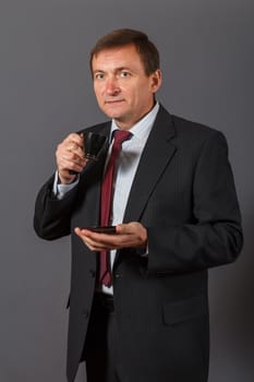 Confident and friendly elegant handsome mature businessman standing in front of a grey background in a studio taking a cup of coffee