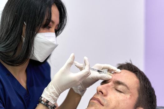 A doctor with a mask injecting collagen into a man's forehead to rejuvenate his face in a clinic