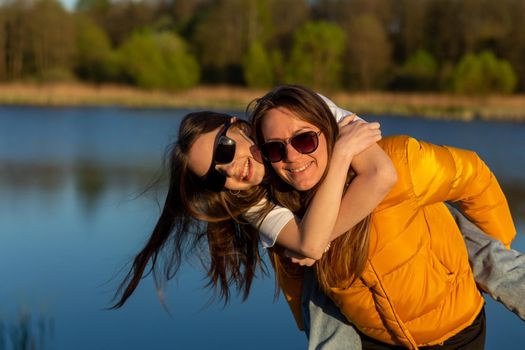 Playful mother giving daughter piggy back ride at spring lake shore. Both laughing and look happy. Spring in lake background. Closeup.