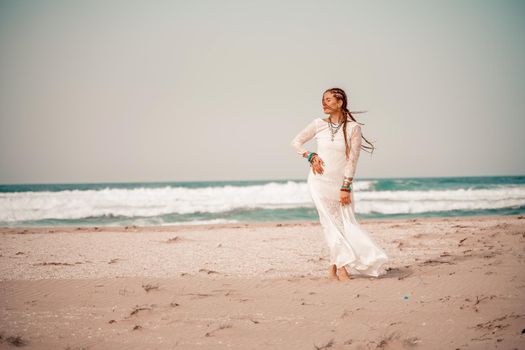 Model in boho style in a white long dress and silver jewelry on the beach. Her hair is braided, and there are many bracelets on her arms