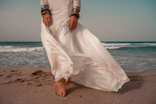 Model in boho style in a white long dress and silver jewelry on the beach. Her hair is braided, and there are many bracelets on her arms