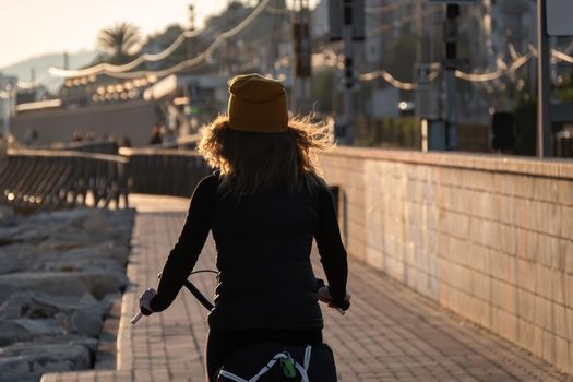 Young girl in a yellow hat with curly hair fluttering in the wind riding a bicycle at sunset on the promenade road of Maresme, Catalonia, Spain.