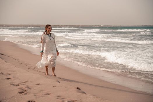 Model in boho style in a white long dress and silver jewelry on the beach. Her hair is braided, and there are many bracelets on her arms