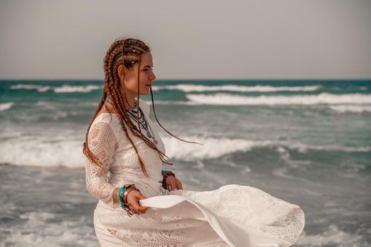 Model in boho style in a white long dress and silver jewelry on the beach. Her hair is braided, and there are many bracelets on her arms