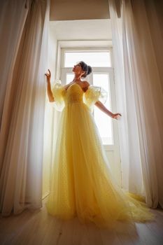 A woman's silhouette in a golden luxurious dress against the background of a window holds a curtain with her hands. Elegant lady in a yellow long silk dress with bare back, back view