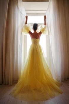 A woman's silhouette in a golden luxurious dress against the background of a window holds a curtain with her hands. Elegant lady in a yellow long silk dress with bare back, back view