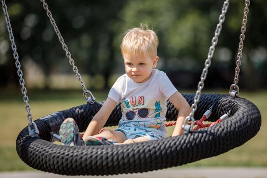 Little boy in striped t-shirt swinging in the swing-nest at a park on summer day. Entertainment for kids outdoors. Street activities concept.