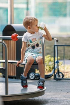 Little boy rides carousel in playground outside. Cute little child. Concept of kindergarten and childhood. Metal carousel for children in summer in the park. Vertical.