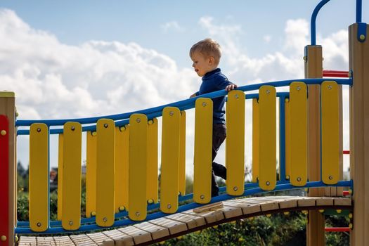 The little boy concentrates walking along the curved bridge of the playground. Curved yellow fence, light blue sky with white clouds in the background.