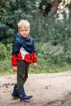 Portrait of a blonde cute boy posing in the nature of autumn. The child wears a colorful waterproof jacket and rubber boots.