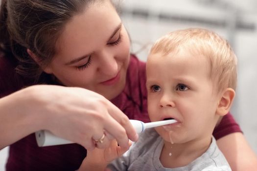 Mother teaching and helping child son how to brush his teeth. The child is not very happy about that.
