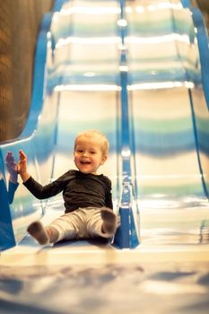 Little kid riding from childrens slides in game center. Happy little kid, children riding up, down on slide, in game center, amuse.