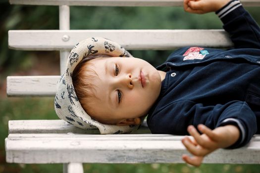 A little cute boy in a hat, lying on a white bench in the garden to rest