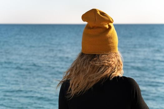 A girl with blond curly hair in black sport jacket and yellow cap is looking at the blue sea. Copy space. View from the back.