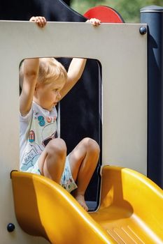 A little boy is ready to slide on a yellow, outdoor playground plastic slide. Vertical.