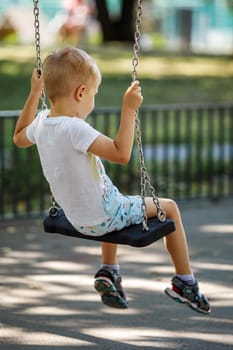 A blond boy in a white T-shirt swings on a swing in the park on a summer day, photo from behind.