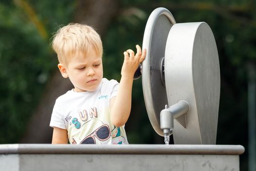 A happy little boy plays with a water tap in a city park. Special water equipment for children's games on a hot summer day outdoors. Horizontal photo.