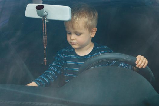 Cute little boy driving fathers car. Portrait of a child sitting in a car behind the wheel from the front. The boy plays and imagines he is driving, he controls the radio with the other hand.