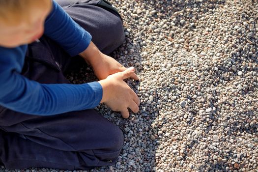The little boy kneels on the gravel, he touches the small pebbles with his hands. The child becomes acquainted with the environment and materials.