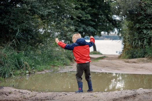 Little boy playing in rainy summer park. Child with waterproof coat and boots jumping in puddle and mud in the rain. Outdoor fun by any weather. Happy childhood.