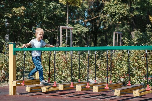 A brave little boy goes across the bridge of the monkeys in a city playground, against a green foliage background.