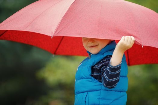 Happy child with an umbrella playing out in the rain in the summer outdoors. The boy smiles and hides his face under an umbrella.