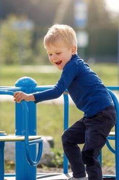 Child on merry-go-round. Boy spins on carousel for children on playground. Junior schoolboy plays alone. Child in blue sweater with blond hair. Guy is holding on tight to handrail.