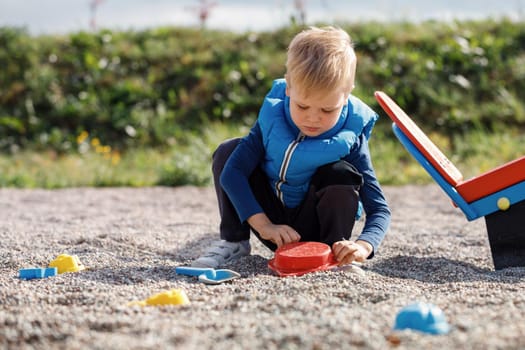 A little boy with a blue vest plays outdoors with small pebbles and colorful toys. Leisure time in the fresh air, a healthy child concentrates on playing.
