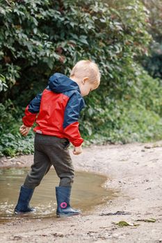 Child in a red waterproof coat in the rain jumping in puddles. A boy stands with his head bowed, a bright raincoat. Kid playing autumn park. Outdoor fun by any weather.