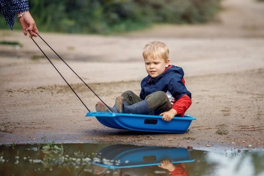 A little boy sitting on a sled skates in the mud near the swamp. Summer time after the rain, sled towing his mom they joke.