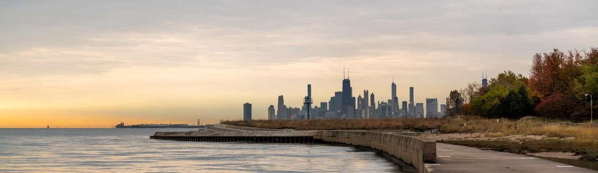 A panorama of the Chicago Skyline at sunrise with orange clouds in the sky and water below in autumn with colorful tree foliage.