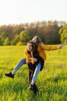 Playful mother giving daughter piggy back ride at green field. Both laughing and look happy. Spring in forest background. Closeup.