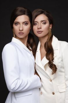 Portrait of two beautiful sisters with elegant make up. Brunette twins with long hair standing close to each other and looking seriously. Young models with big earrings posing together at camera.