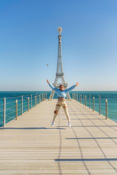 Large model of the Eiffel Tower on the beach. A woman walks along the pier towards the tower, wearing a blue jacket and white jeans