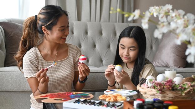 Smiling asian mother and her daughter enjoy painting Easter eggs together in living room.