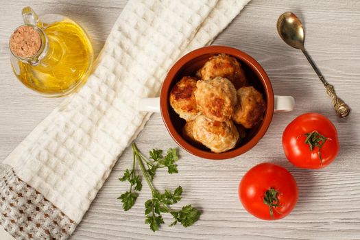 Fried meat cutlets in ceramic soup bowl, red tomatoes, metal spoon, branch of fresh parsley, kitchen towel and glass bottle with sunflower oil on grey wooden boards. Top view