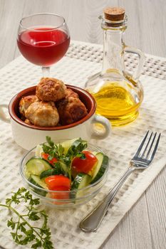 Fried meat cutlets in ceramic soup bowl, glass of red wine, bottle with sunflower oil and salad of fresh cucumbers and tomatoes in glass bowl on white kitchen towel