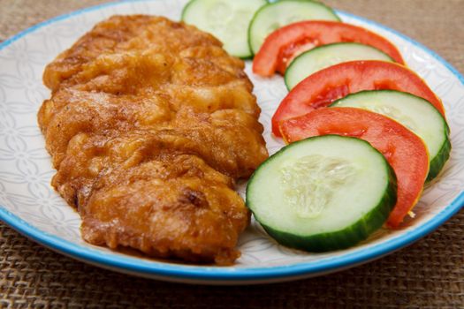 Plate with fried meat chop in batter and sliced tomato and cucumber on plate