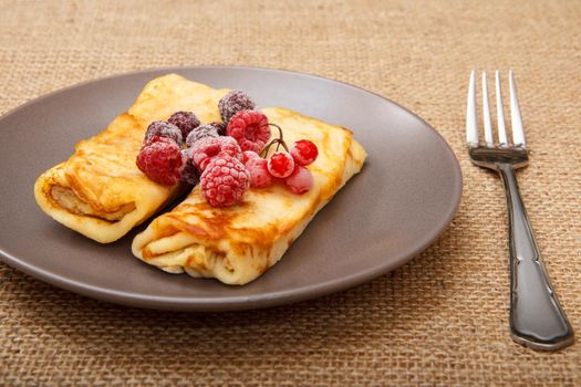 Homemade pancakes filled with cottage cheese and topped with frozen raspberries and blackberries on plate and fork on sackcloth background