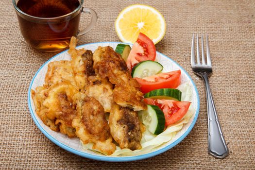 Plate with pieces of fried fish and freshly sliced tomatoes and cucumbers, cup of black tea with slice of lemon, fork on sackcloth