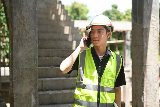 Young engineer builder in helmet talking on mobile phone while standing at construction site.